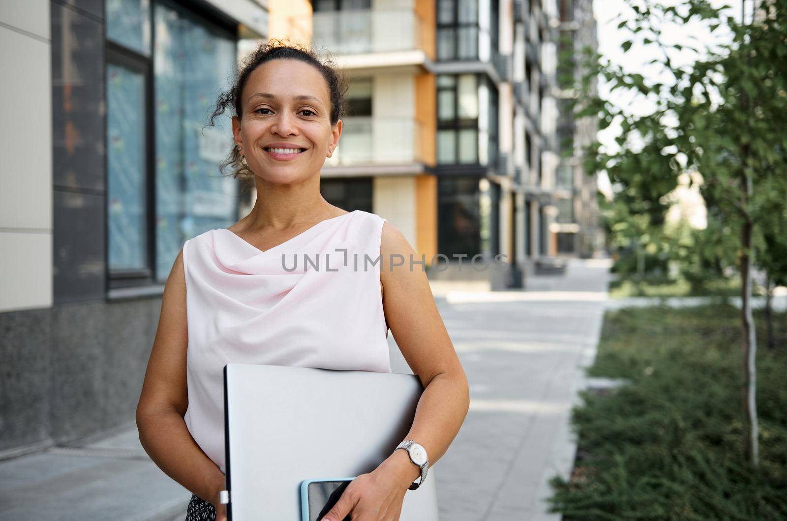 Happy brunette mixed race woman in casual attire holding laptop computer, smiling looking at camera on the tall buildings background. Business, freelance, office work concept by artgf