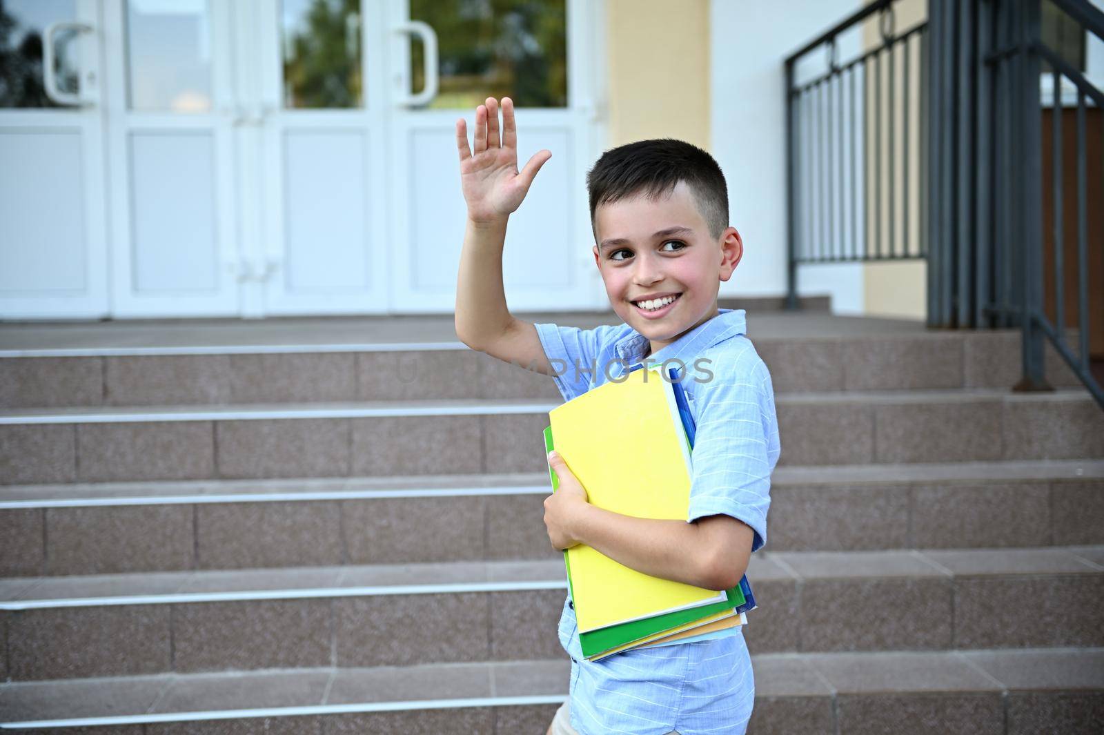 Handsome elementary student, pupil, coming back to school. Adorable schoolboy holding a workbooks and waving hello on school establishment background.