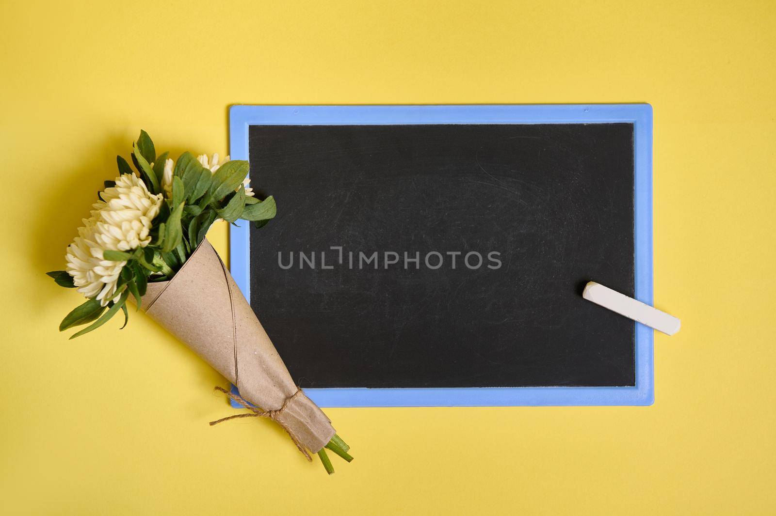 Flat lay composition of delicate beautiful stylish bouquet of asters flowers in craft wrapping paper tied with a rope on a blank empty chalkboard with space for text isolated on yellow background by artgf