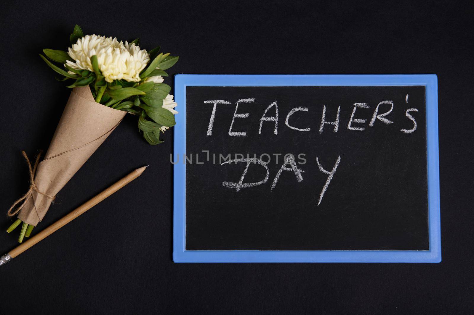 A wooden pencil and beautiful bouquet of asters flowers in craft wrapping paper lying down next to a chalk board with lettering Teacher's Day. Flat lay on black background with copy space