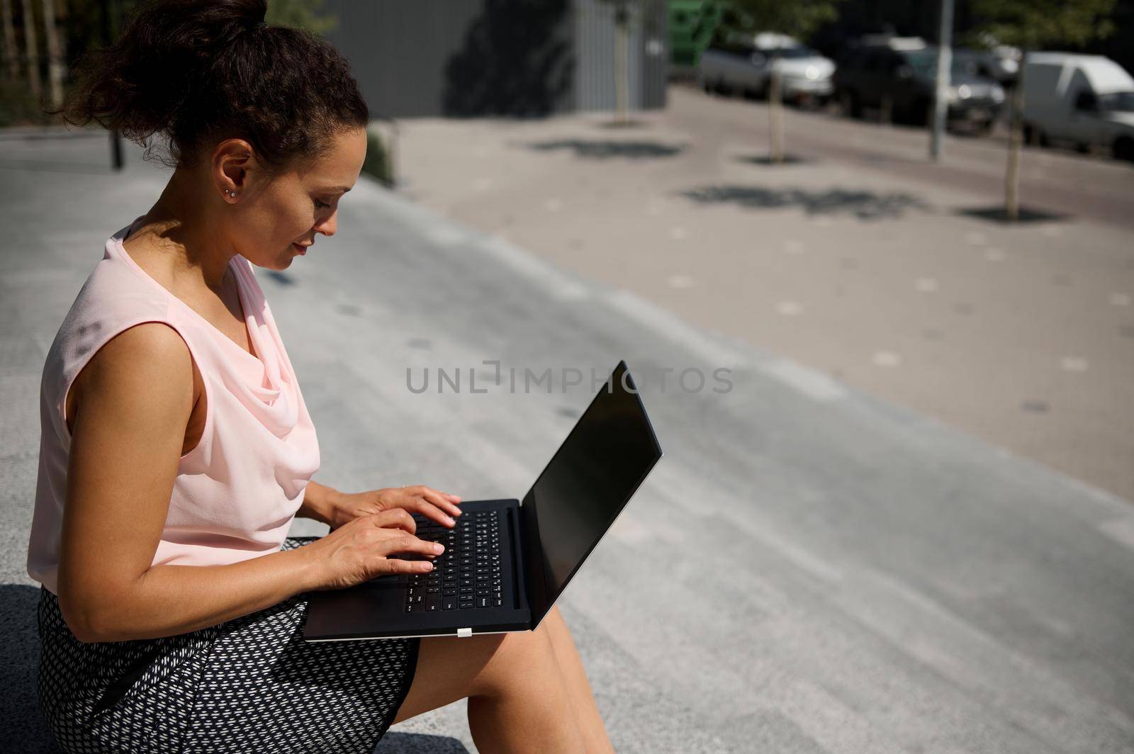 Side portrait of busy freelance woman, start-uper , office worker using laptop sitting on steps during coffee break. Office worker on lunch break in urban background by artgf