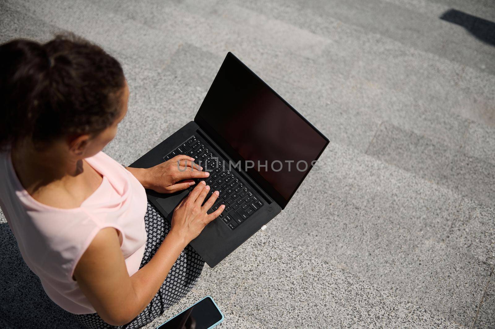 High angle view of a woman sitting on steps and typing text on on laptop with blank black monitor screen with copy space for advertising on the urban background. Business concept