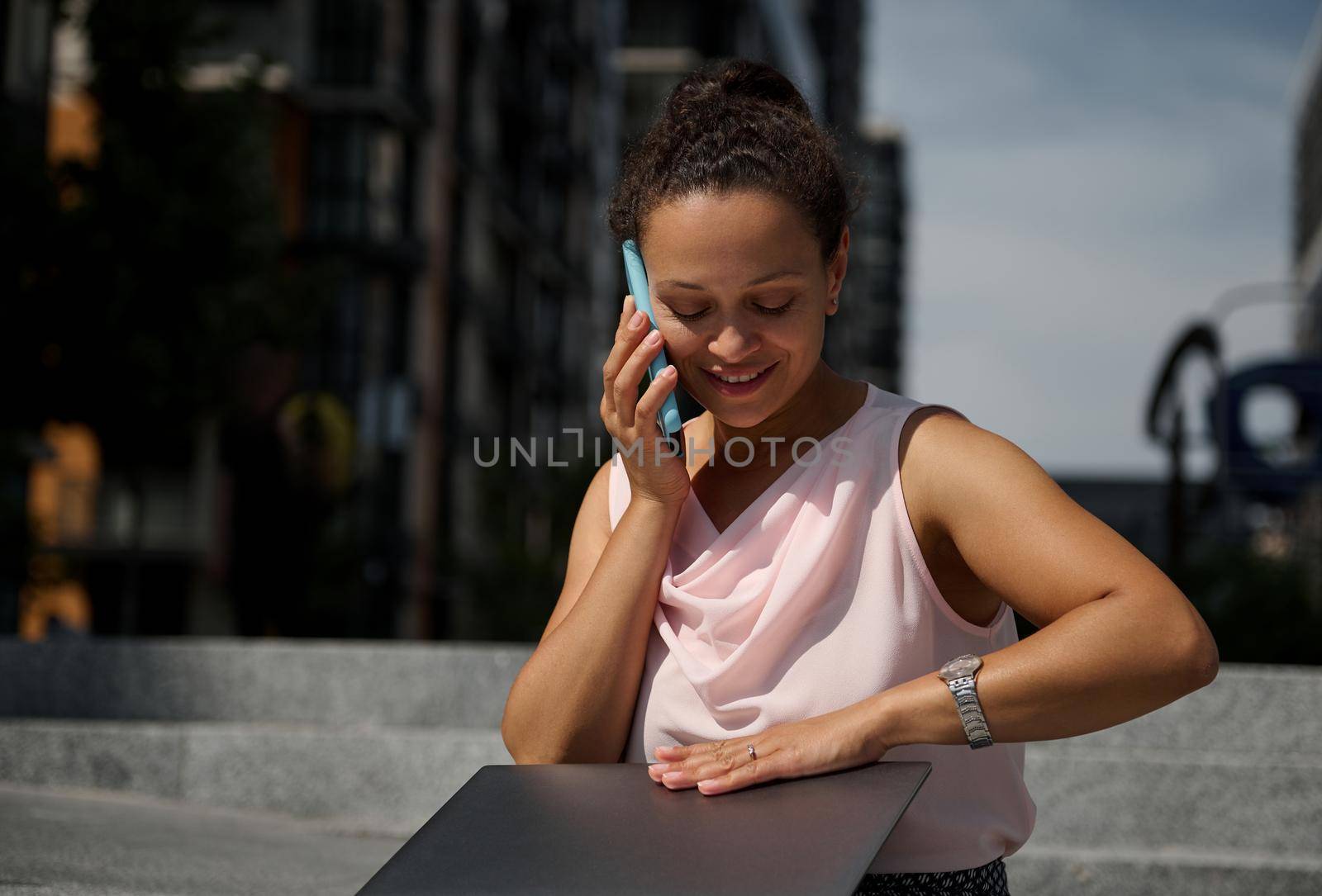 Smiling African American freelance woman talking on mobile phone, using laptop while sitting on steps in urban background. Freelance, business, e-learning, online meeting and distant work concept