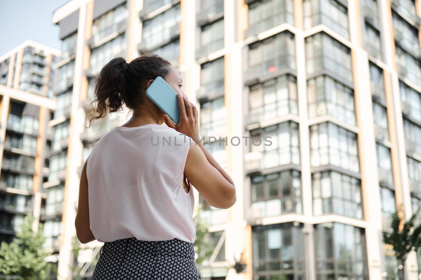 View from the back of a young African American mixed race woman talking on mobile phone on the background of urban high buildings. Business and communication concepts by artgf