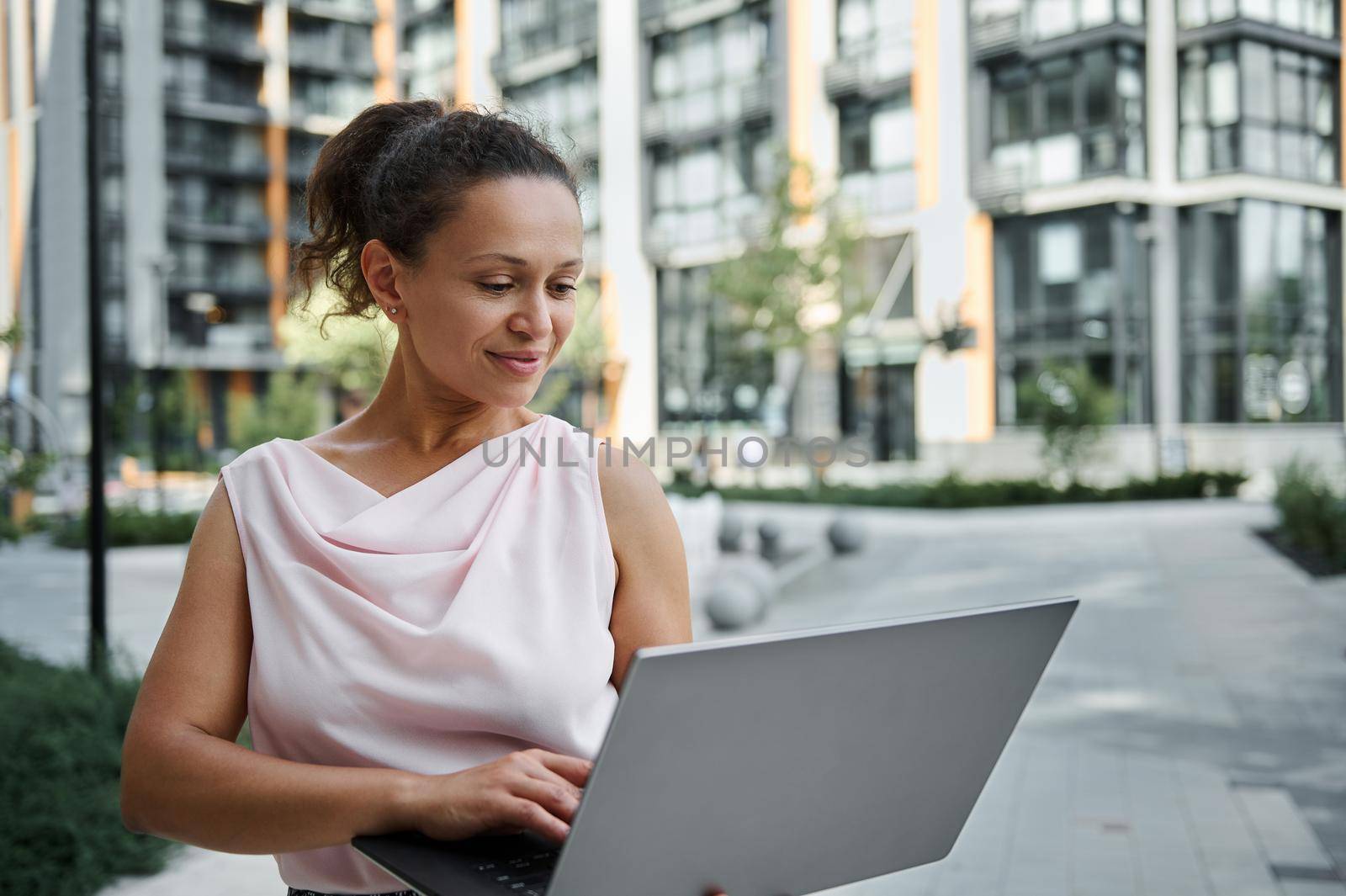 Attractive Hispanic young woman working on laptop, cutely smiling with toothy smile while looking at the screen monitor, standing on the urban buildings background. Business and communication concept by artgf
