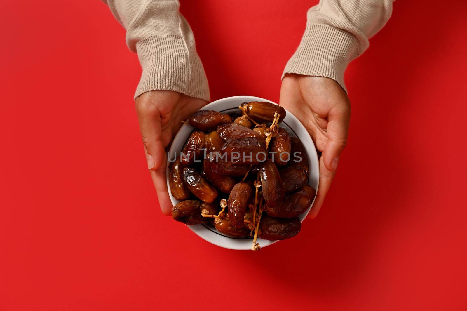 Flat lay composition of hands holding a delicious and sweet plate full of fresh dates, isolated on red background with copy space