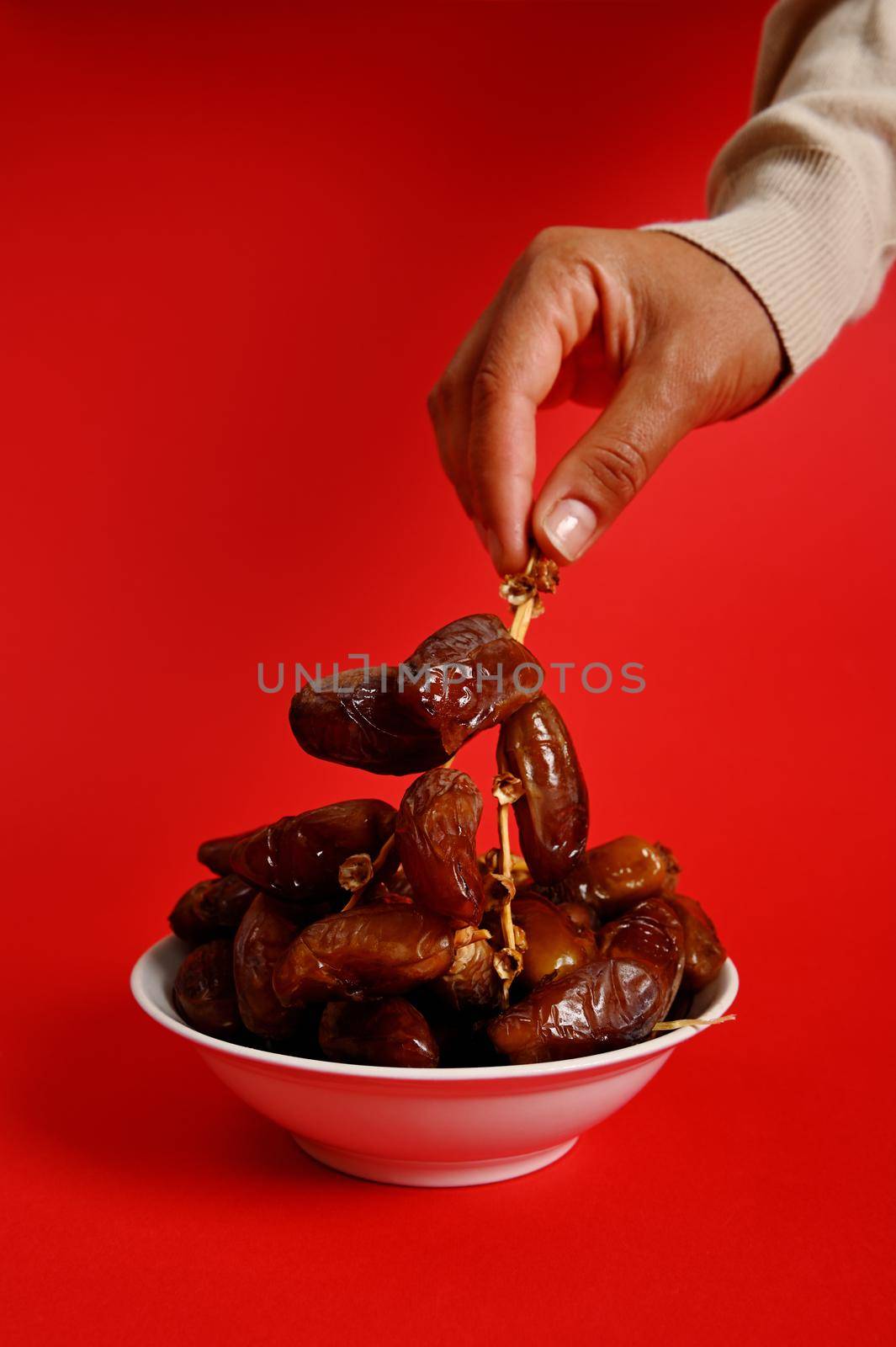 Female hands holding a branch of fresh tasty sweet dates. Vertical shot on red background with copy space. Dates for breakfast after interruption and fasting in Ramadan