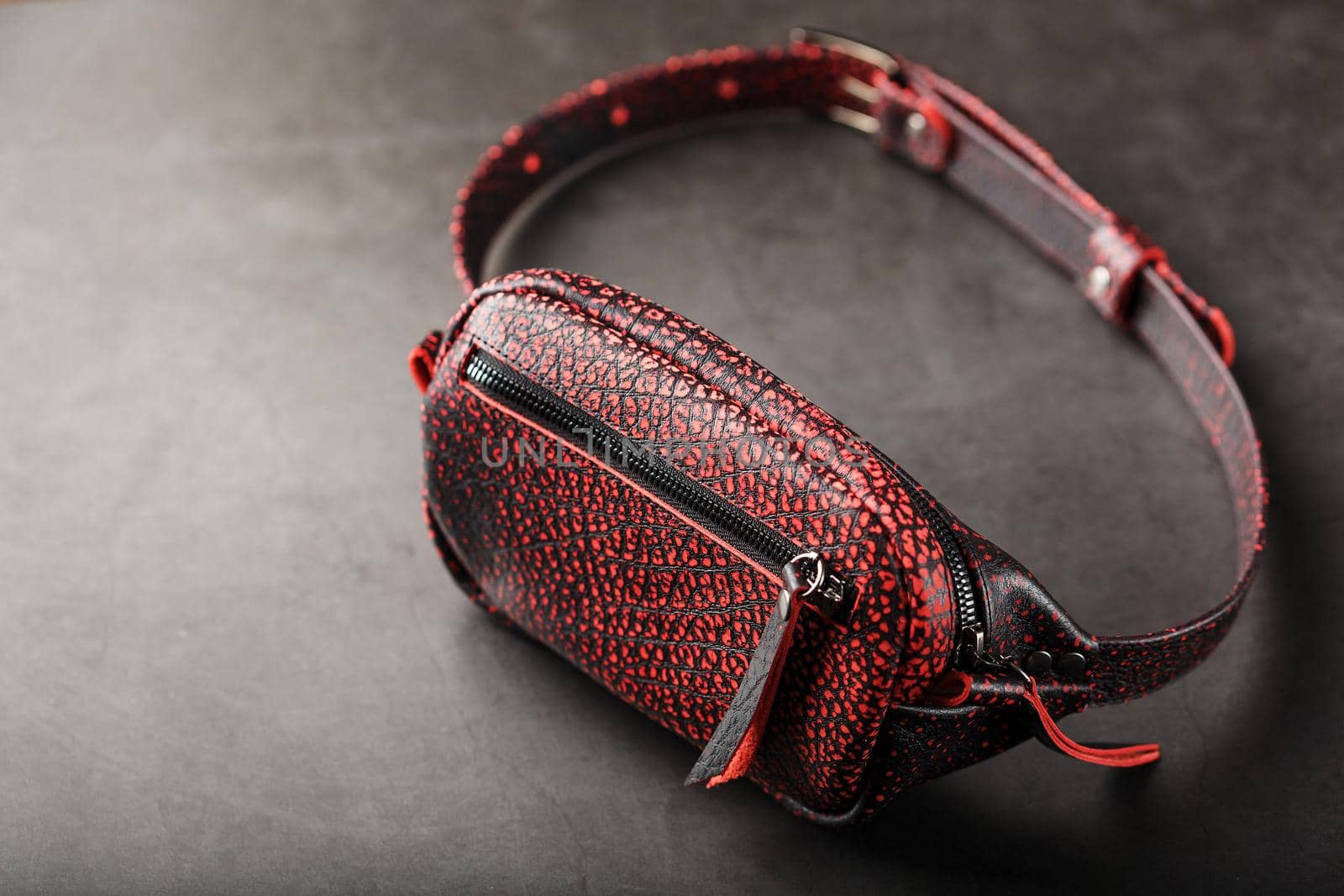 Belt bag made of textured, spotty black and red leather on a stone dark background. Elegant black and red bag with a zipper. Genuine leather, handmade. Banana bag