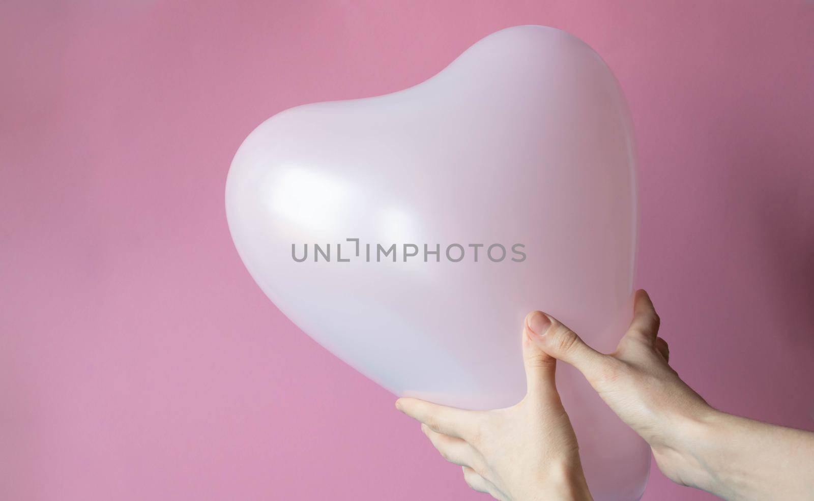 Women's hands hold a pink ball in the shape of a heart, highlighted on a pink background. by lapushka62