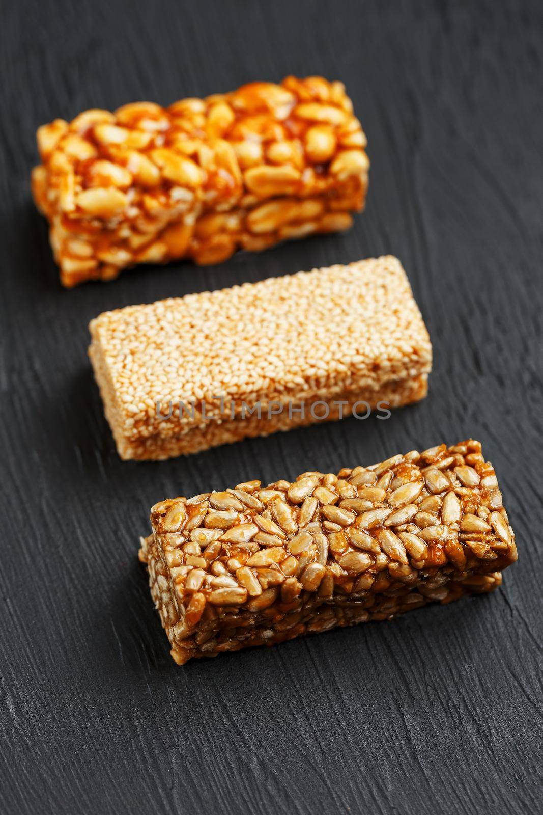 Cereal granola bar with peanuts, sesame and sunflower seeds on a cutting board on a dark stone table. View from above. Granola bar is a creeping and sweet snack. Three Assorted Bars
