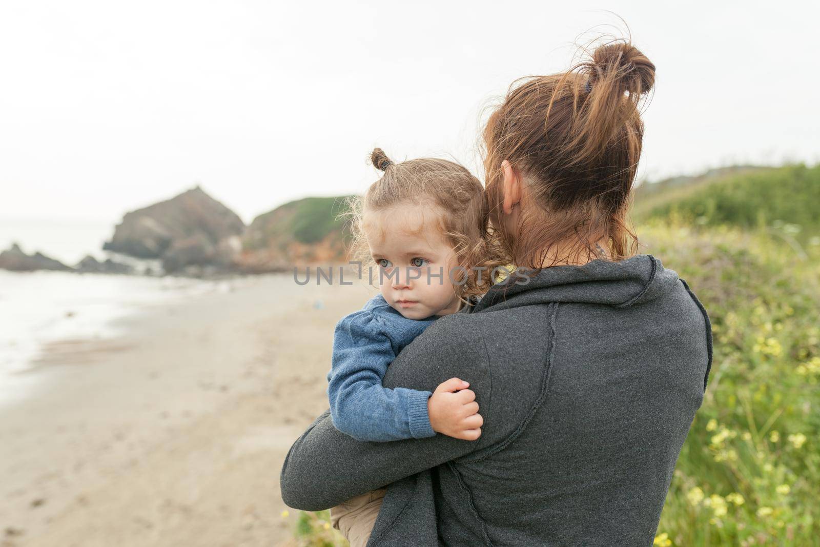 Mother and little daughter standing rock ocean beach autumn season. Caucasian family with one child traveling together. Mom holding little girl 2 years on hand Sea landscape background