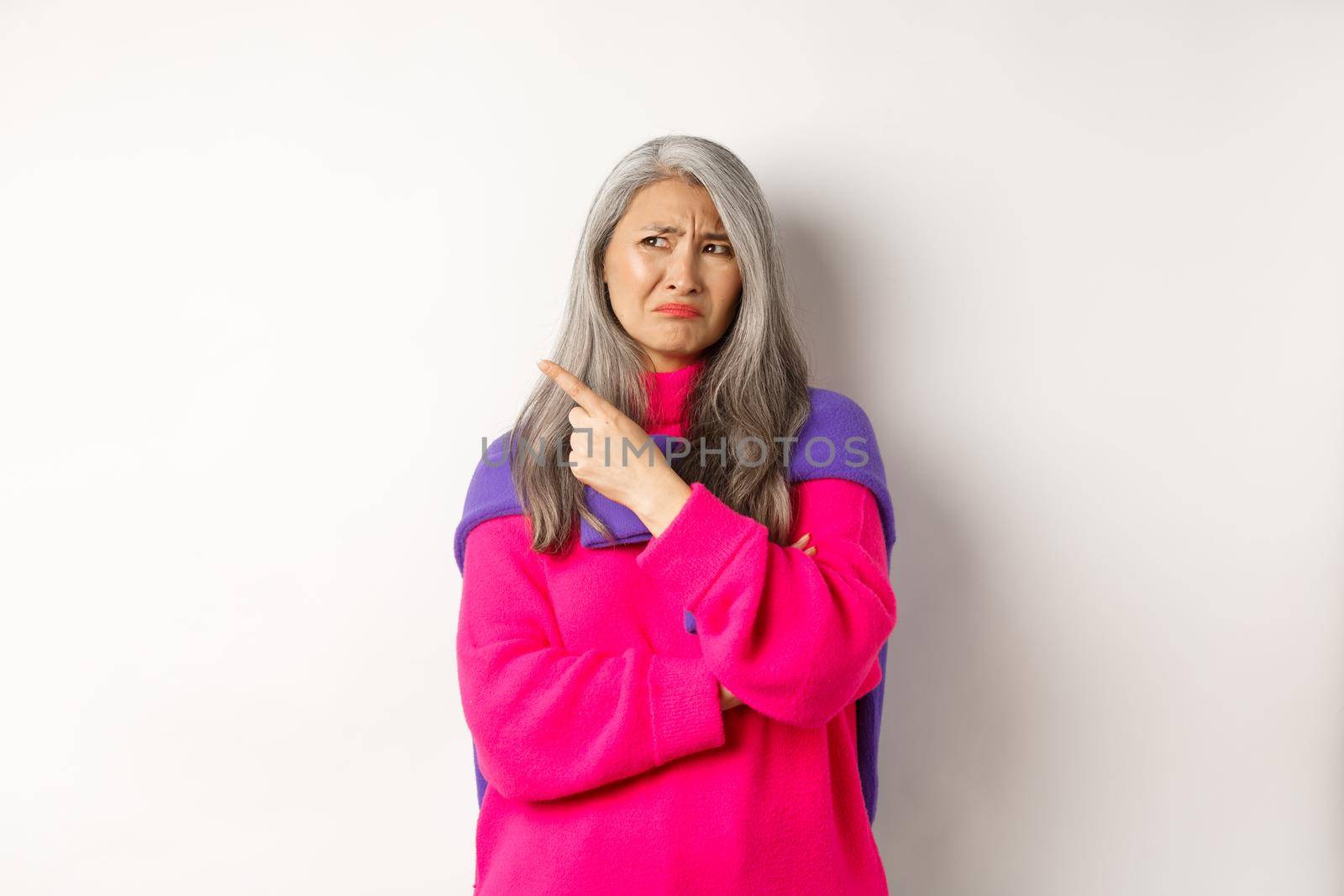 Disappointed asian grandmother grimacing disgusted, pointing finger left and complaining on something bad, standing over white background.