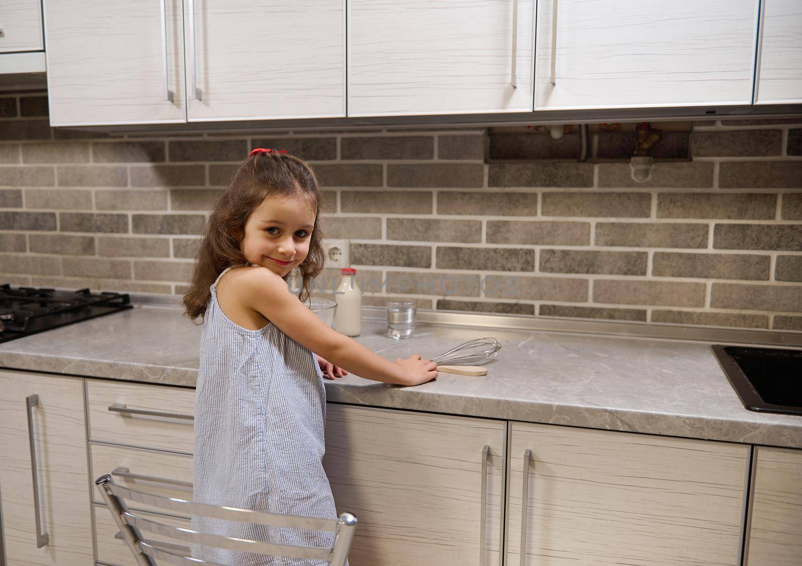 Adorable Caucasian baby girl standing on a chair by kitcheb countertop and smiles looking at camera through her shoulders by artgf