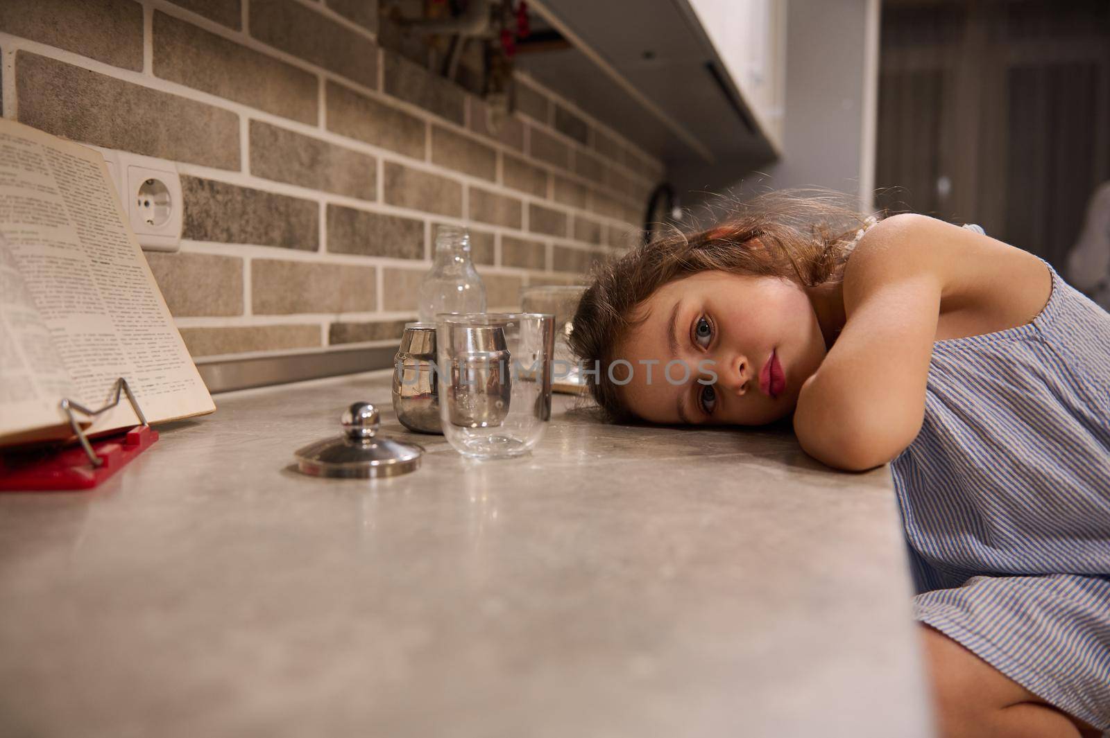 Portrait of an adorable beautiful Caucasian 4 years old little girl in a blue dress looking at the camera while putting her head on the kitchen countertop while making dough