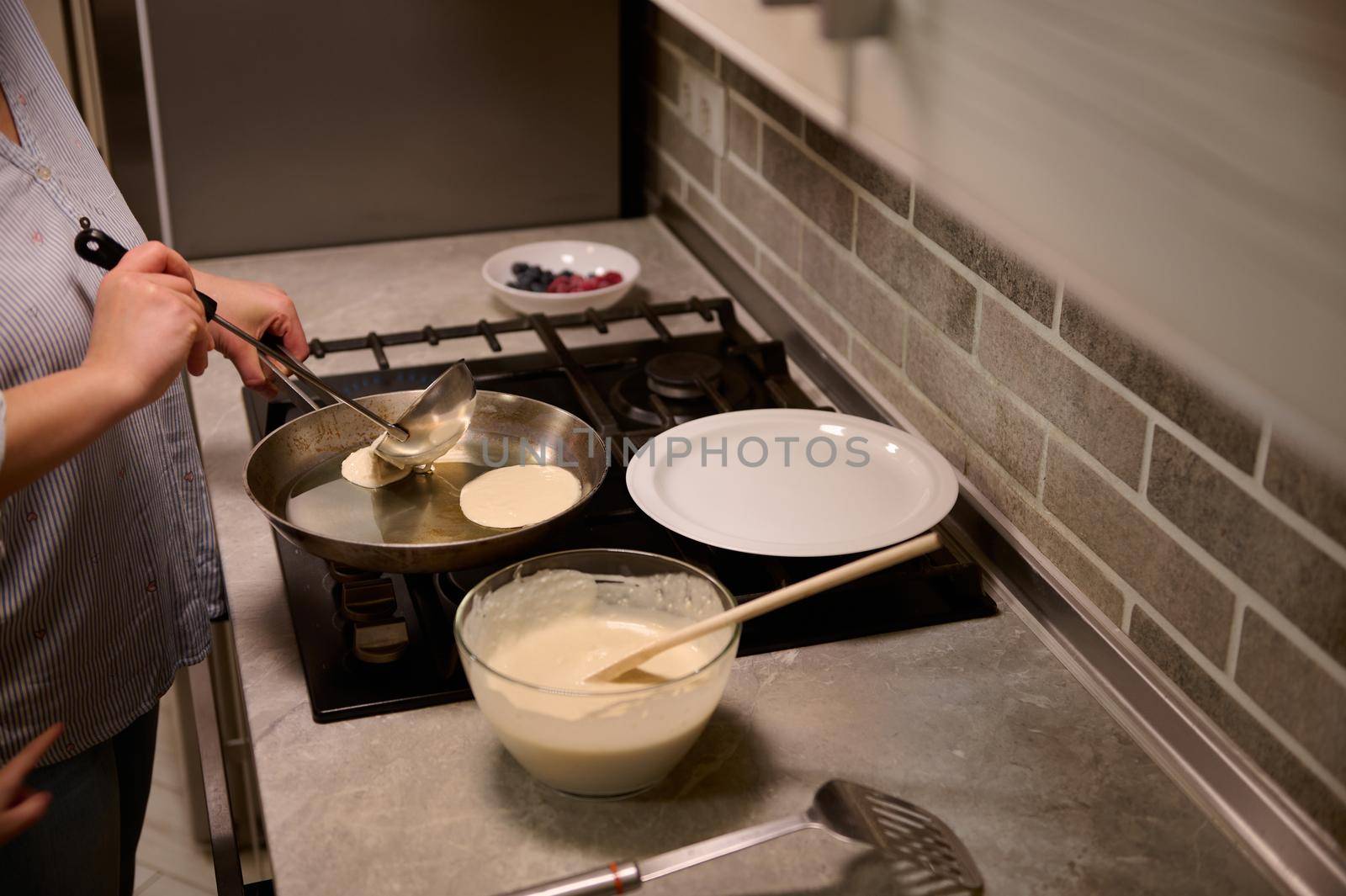 Close-up of a chef woman, housewife pouring round-shaped liquid dough and cooking pancakes in a frying pan, smiles sweetly, standing near the black stove in the home kitchen. Shrove Tuesday concept by artgf