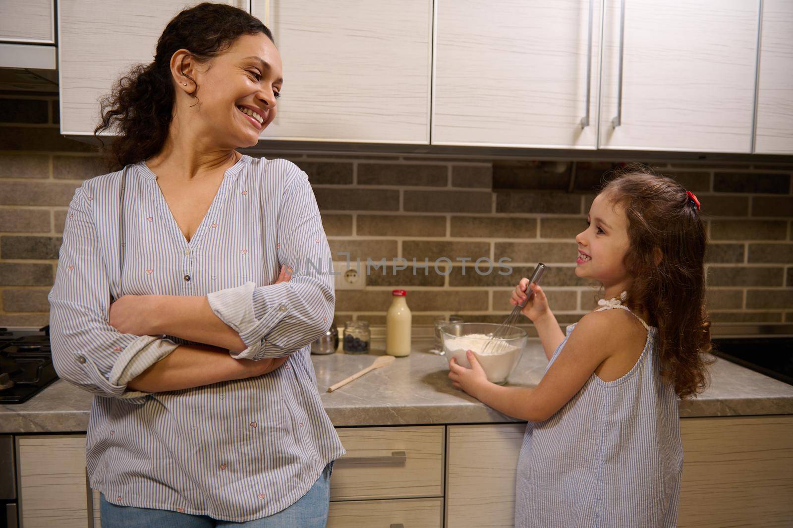 Beautiful smiling woman, happy loving mother leaning at kitchen countertop and admiring her adorable daughter cute baby girl mixing ingredients in a glass bowl while learning kneading pancakes dough by artgf