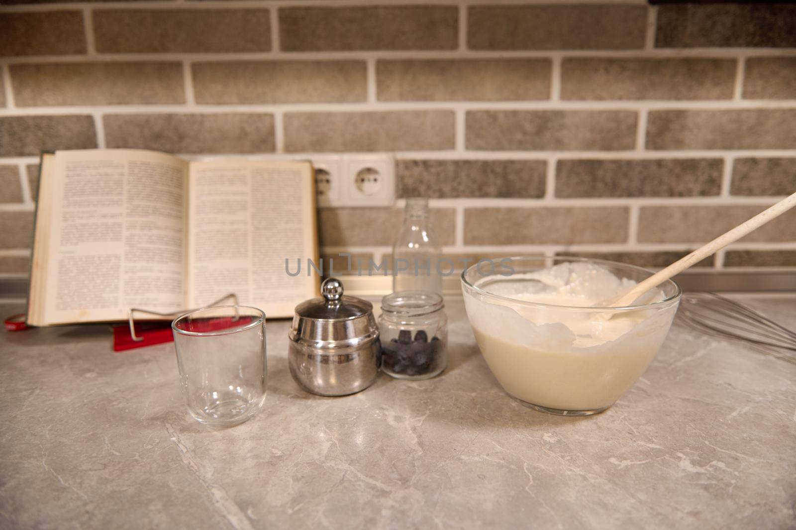 Transparent glass bowl with wooden spoon dropped into a liquid pancake dough, blueberries in a glass container on the background of a recipe-book on a kitchen countertop. by artgf