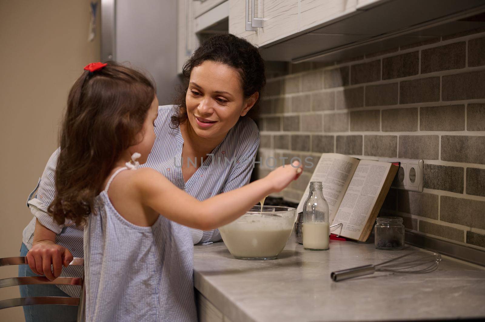 Adorable happy mother smiles to her daughter, cute little girl mixing the ingredients in a bowl while preparing pancake dough. Motherhood, mom teaching her daughter to cook pancakes for Shrove Tuesday by artgf