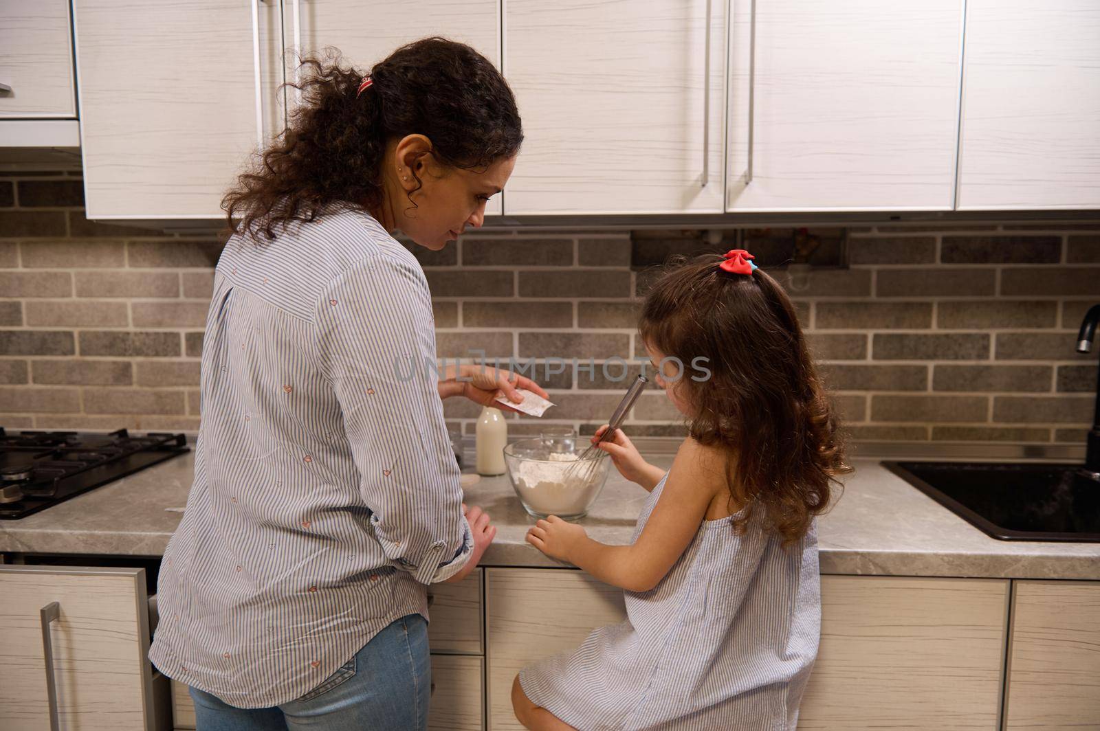 Rear view of charming girl mixing dry ingredients in a glass bowl and her mother pouring some vanilla powder into flour while making pancake dough in home kitchen. Mother and daughter cooking together by artgf