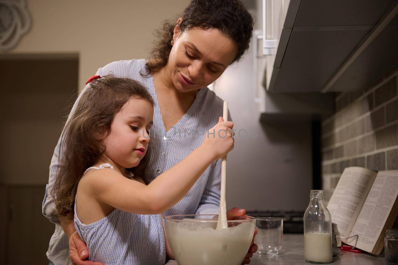 Adorable little girl having fun, concentrated on mixing ingredients and kneading dough in a glass bowl while her loving happy mother teaching her to cook pancake dough. Shrove Tuesday concept by artgf