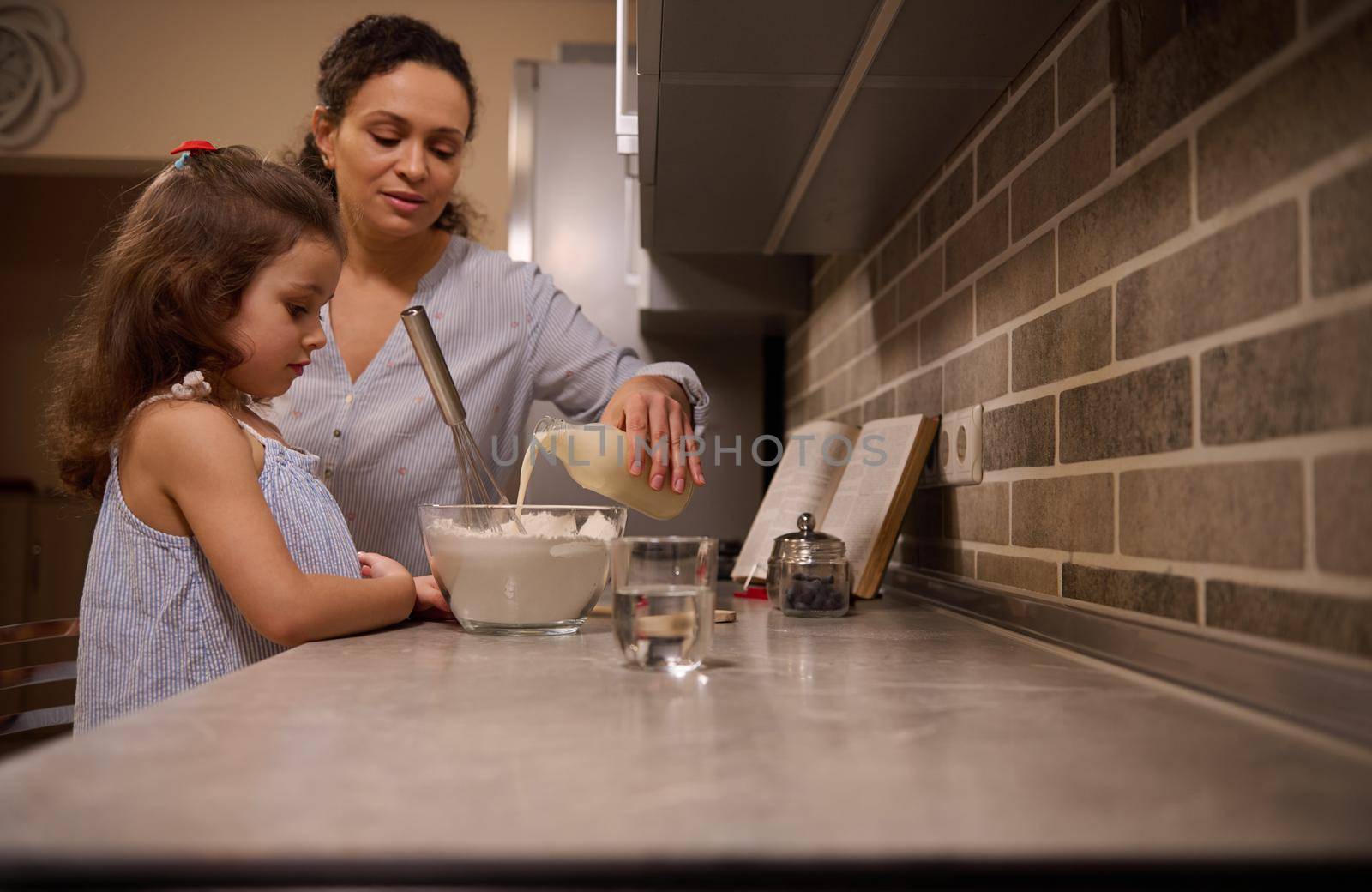 Lifestyle portrait of a mom pouring milk into bowl of flour and mixing ingredients with a whisk to prepare pancake dough. Mother and daughter - beautiful child are cooking together in the home kitchen by artgf