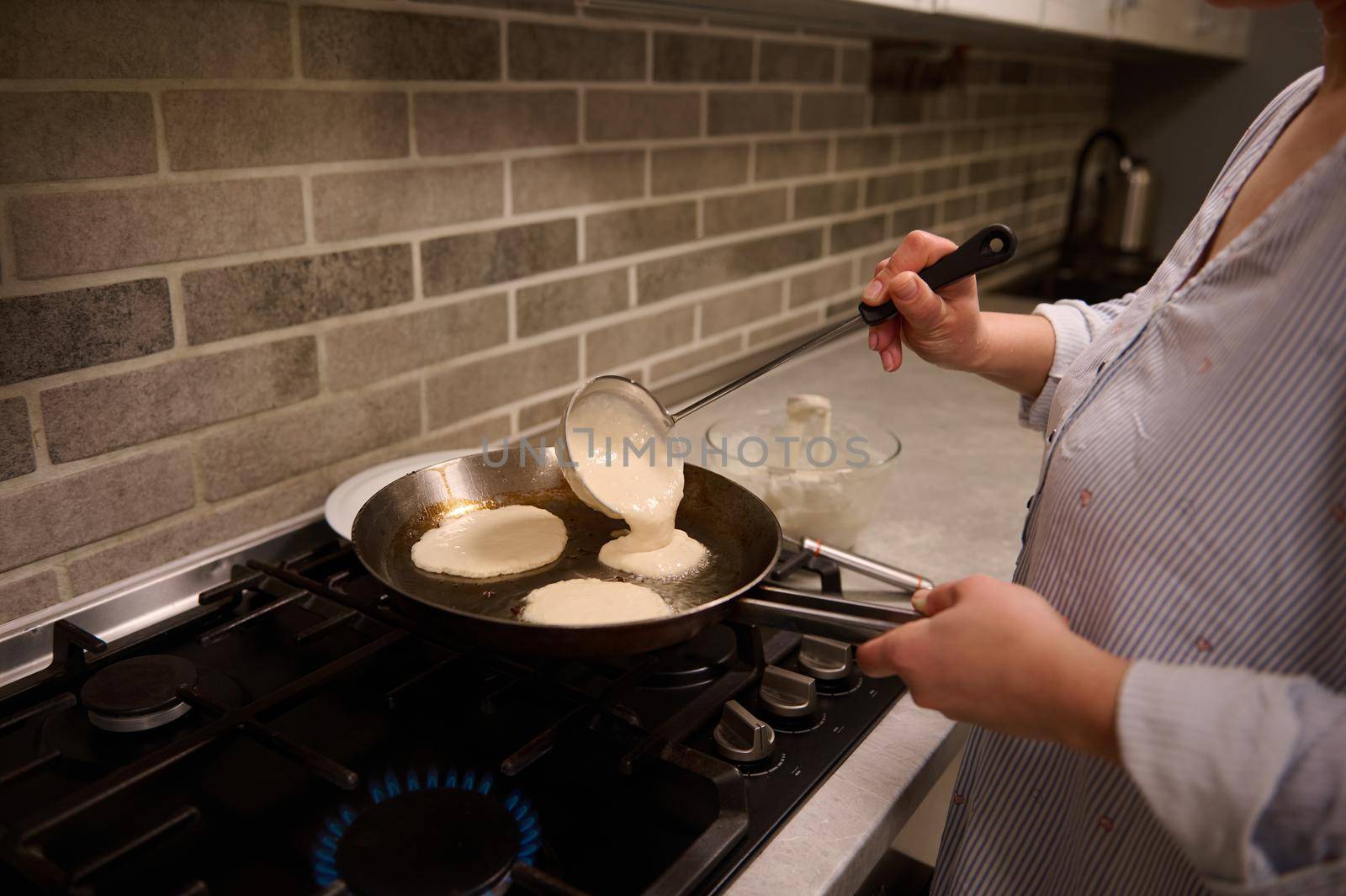 Close-up of a female chef, housewife, pouring round-shaped batter into a frying pan over a black stove and preparing pancakes for Shrovetide. Cooking, Shrove Tuesday, Maslenitsa concept