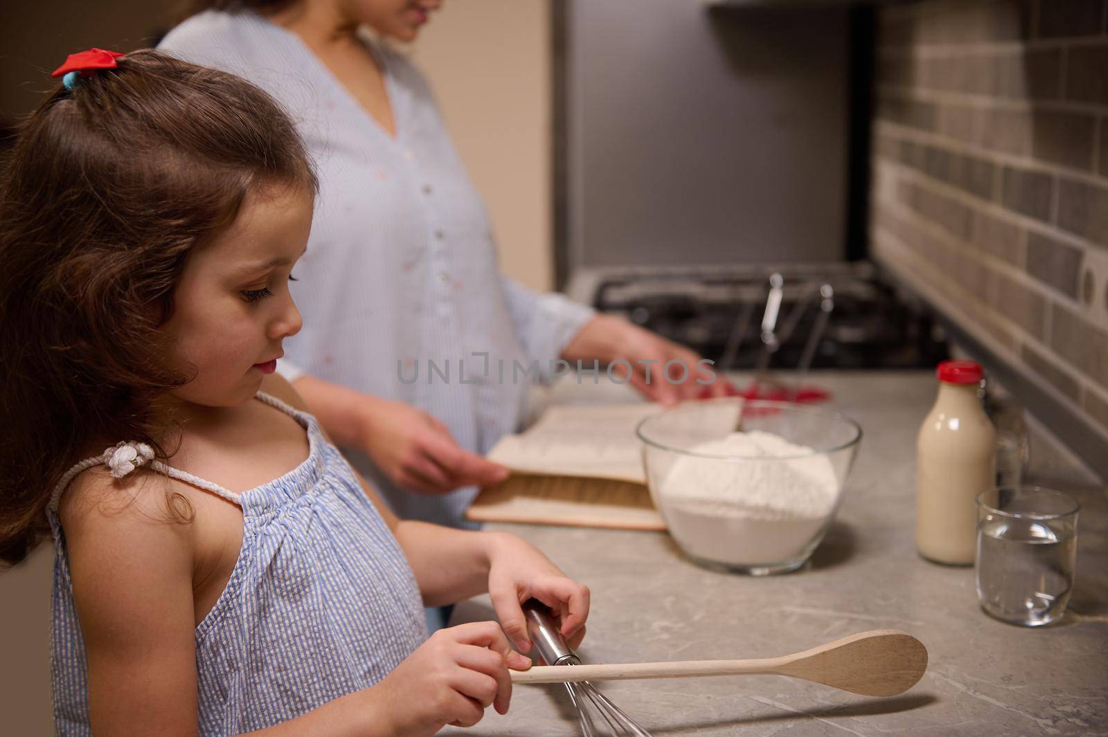 Beautiful child, adorable little girl standing at kitchen counter top, holding a wooden spoon on the blurred background of her mom, searching a recipe on recipe book for preparing pancake dough