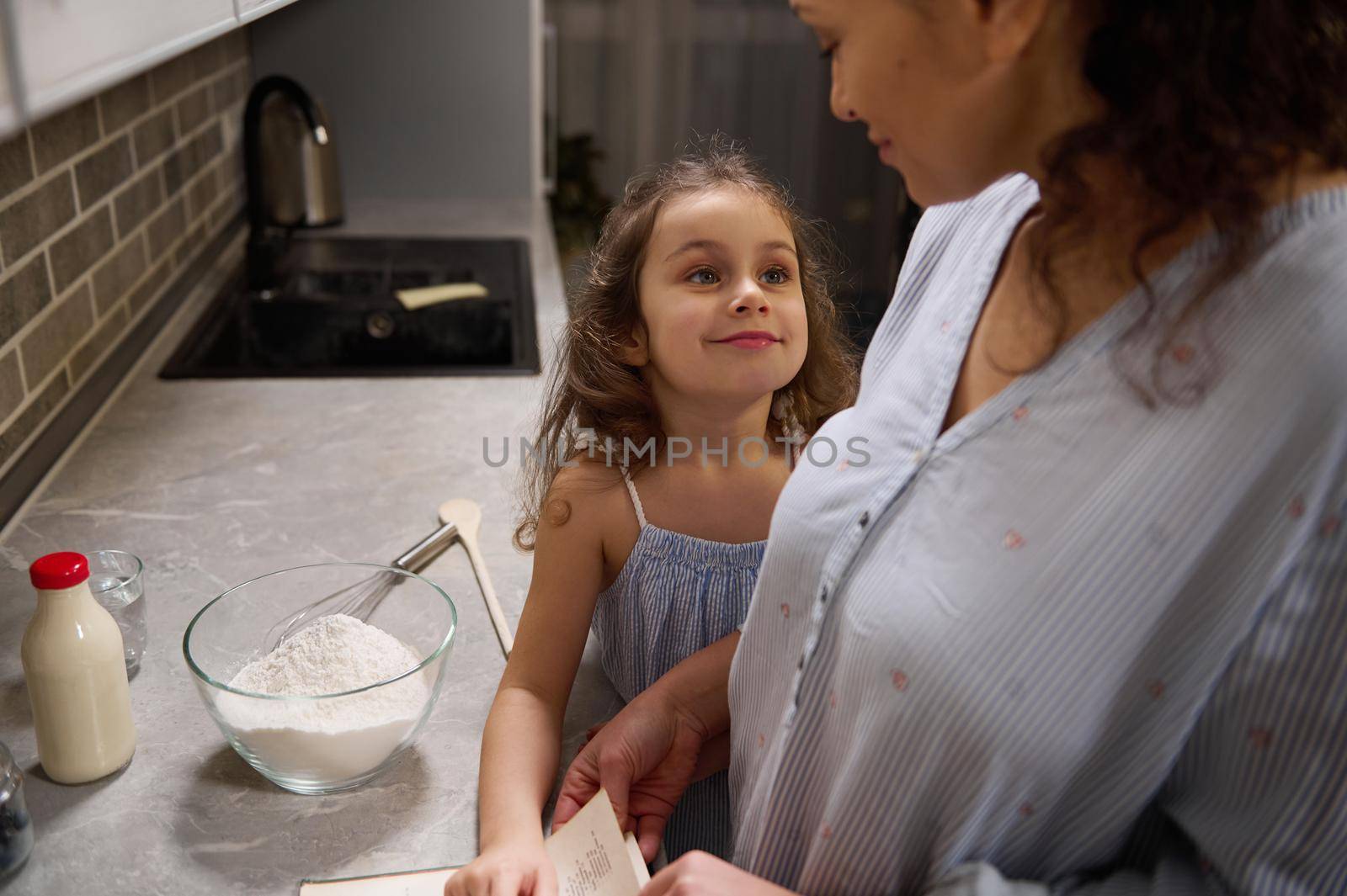 Beautiful happy child, adorable little girl smiles cutely looking at her loving mother, standing at kitchen countertop and cooking together, preparing dough for celebrating Shrove Tuesday by artgf