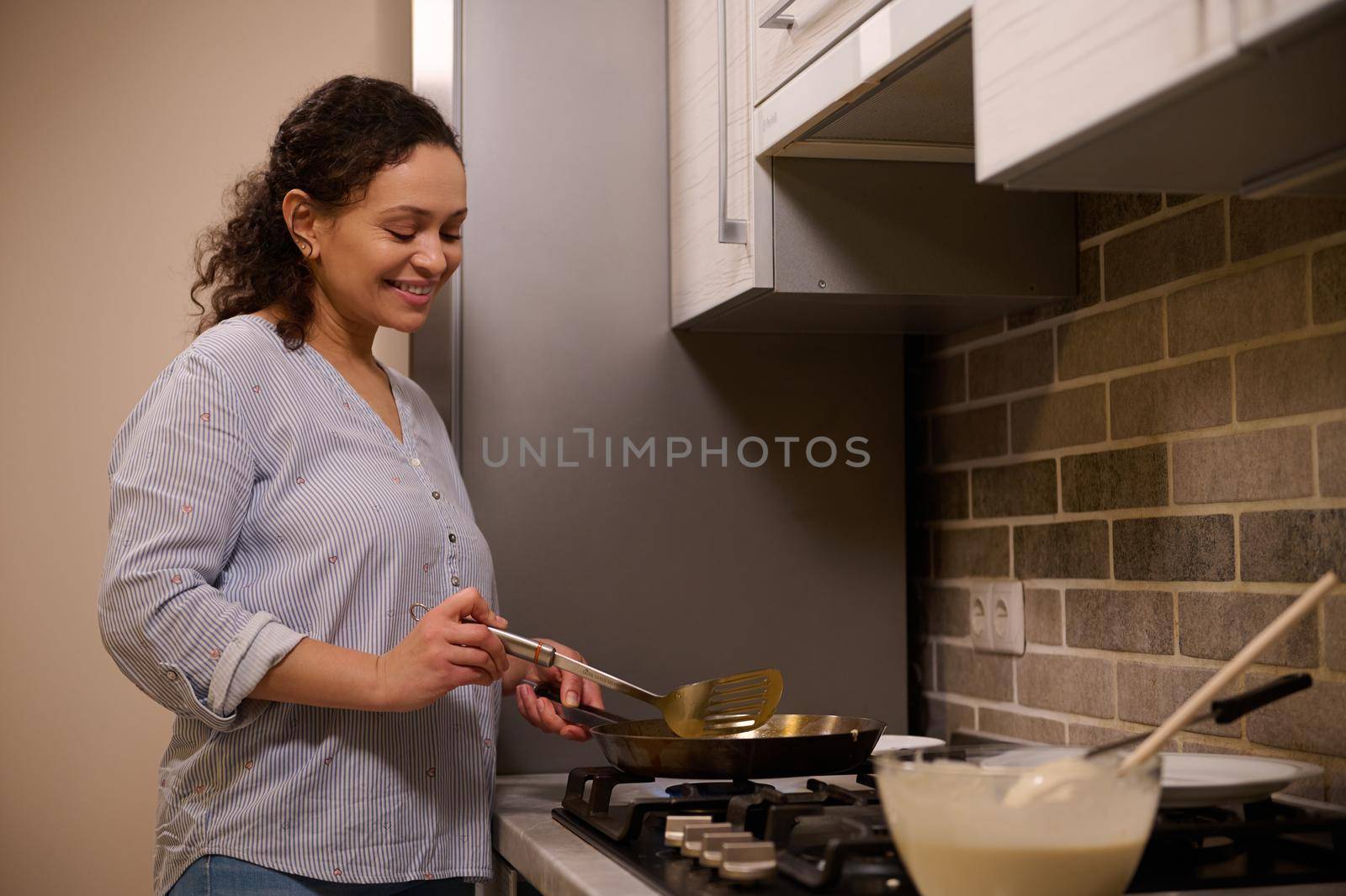 African American woman, happy housewife cooks pancakes in a frying pan, smiles sweetly, standing near the black stove in the home kitchen