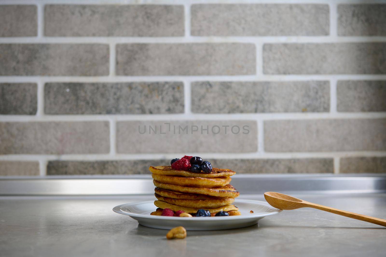 Stack of homemade delicious pancakes garnished with raspberries, blueberries and cashews on a white plate against a gray brick kitchen wall. Shrove Tuesday, Shrovetide concept. Food background