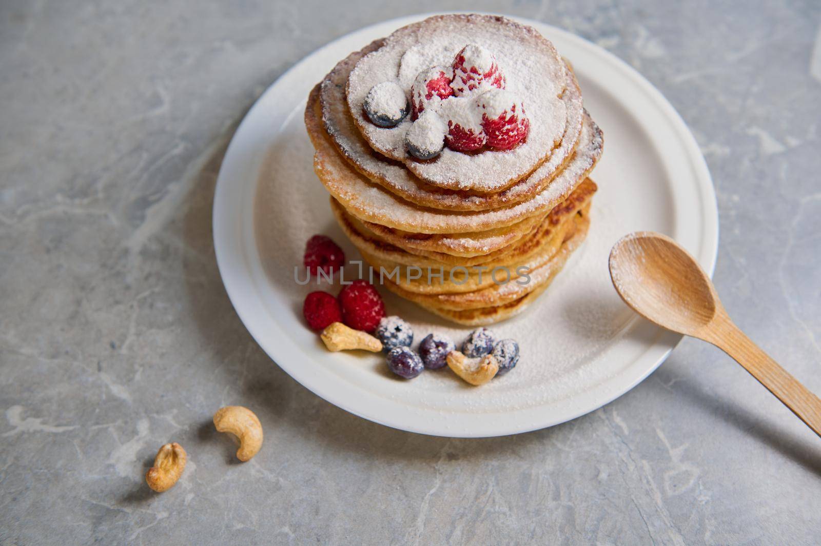 Top view of delicious homemade pancakes sprinkled with powdered sugar and decorated with raspberries, blueberries and cashews served on a white plate with a wooden spoon. Food for Shrove Tuesday