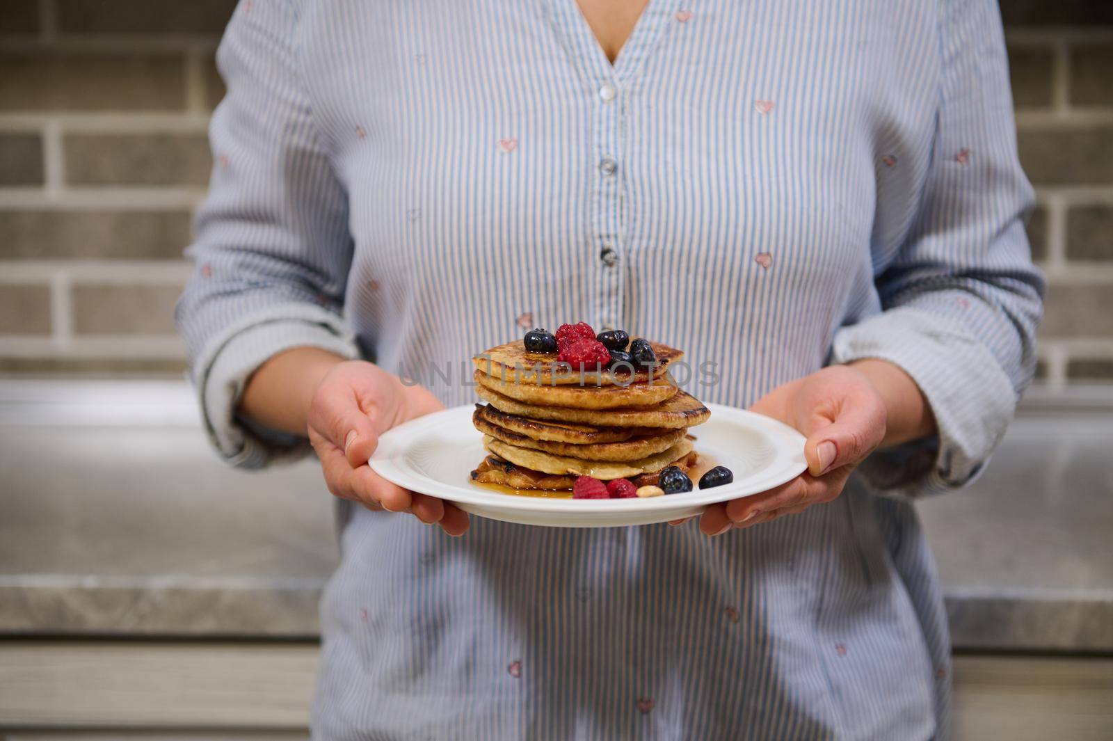 Woman holding plate of delicious homemade pancakes garnished with raspberries, blueberries and cashews, standing against kitchen countertop. Food for Shrovetide, Shrove Tuesday concept. Cropped image. by artgf