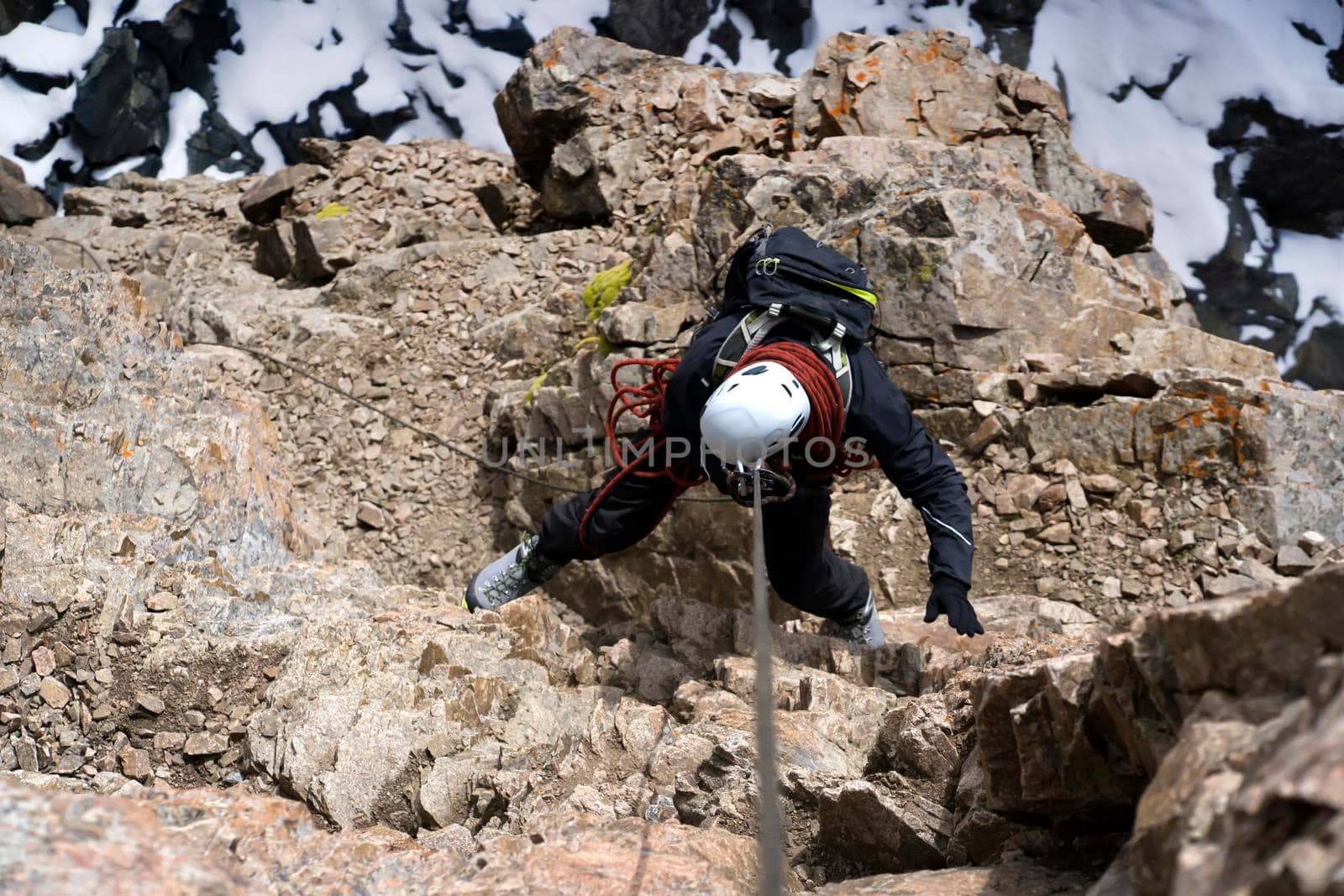 A young man in a helmet and with climbing equipment descends from the top of the mountain on a rope, the climber rappels with aperture-style self-braking belay in the snowy mountains.