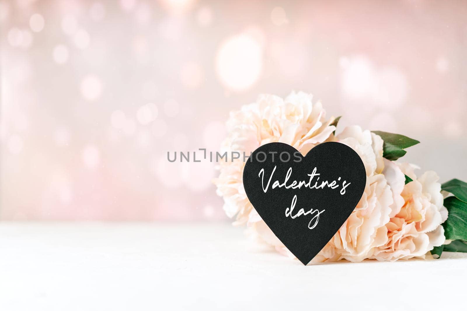Still life Valentines day text festive background with empty black chalkboard heart and flowers on white table. Mockup banner with copy space for design and glowing bokeh