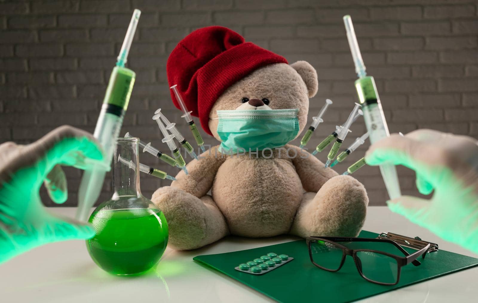 a teddy bear in a medical mask with syringes in his shoulder on the doctor's table is given a second vaccination injection against the corona virus by Rotozey