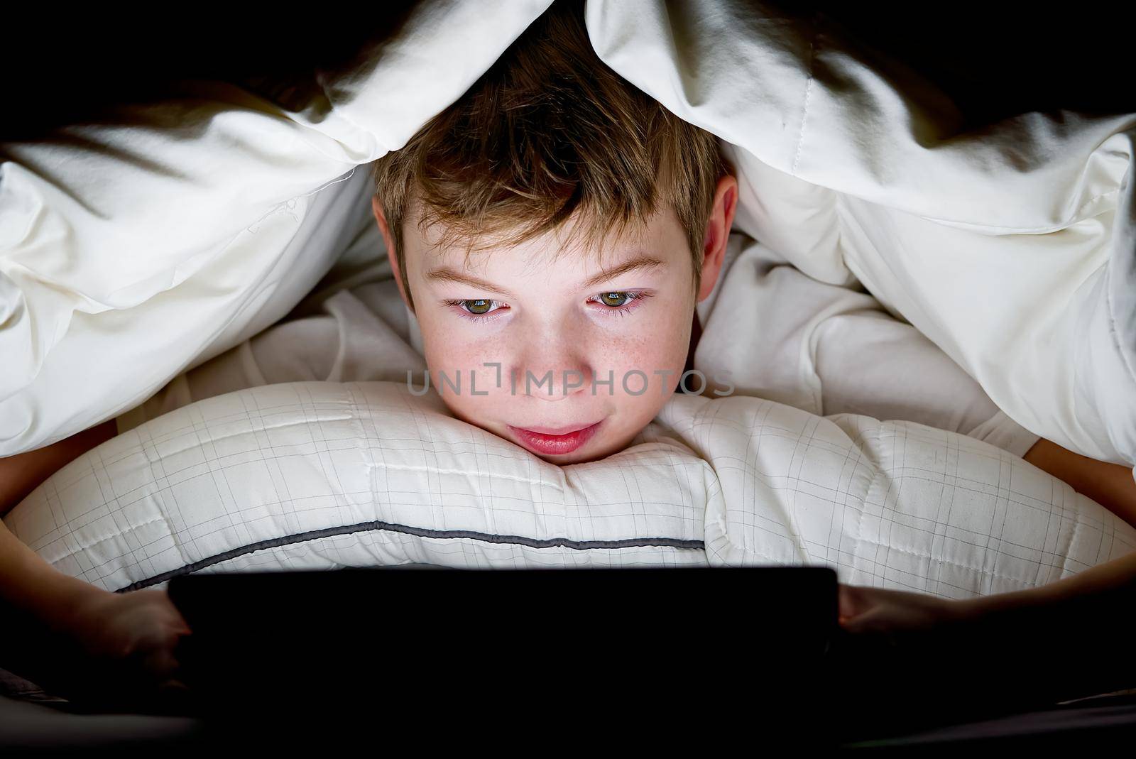 Distance learning. Online education. Happy boy in bed under blanket with tablet in dark. Child's face is illuminated by bright monitor. Quarantine. social media addiction by PhotoTime