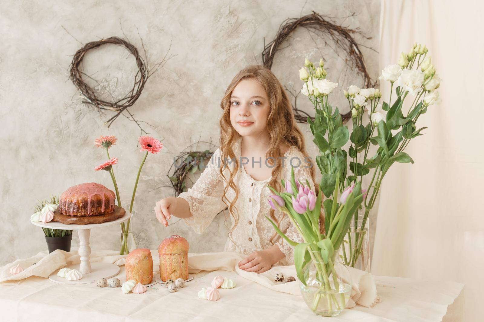 A girl with long hair in a light dress is sitting at the Easter table with cakes, spring flowers and quail eggs. Happy Easter celebration