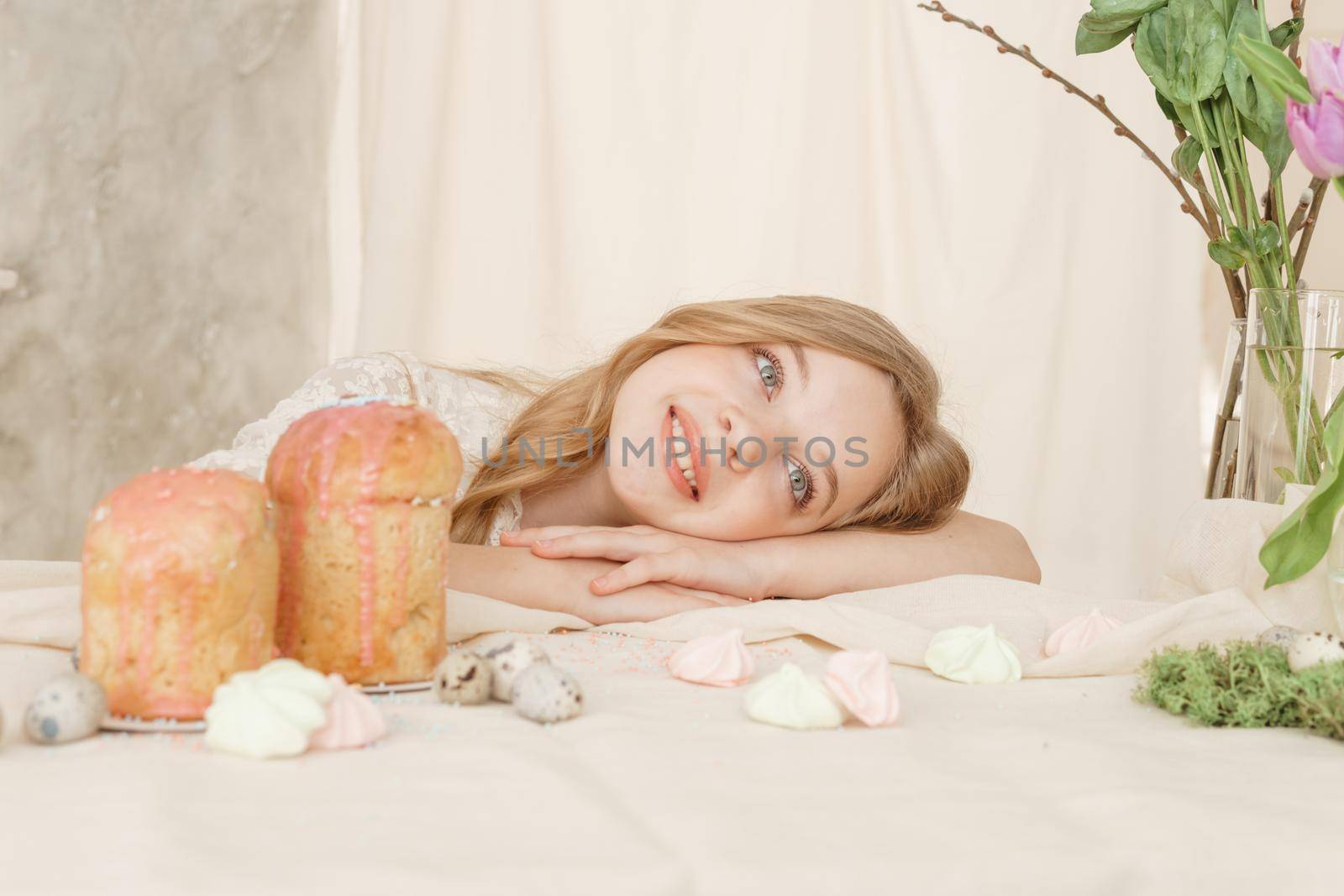 A girl with long hair in a light dress is sitting at the Easter table with cakes, spring flowers and quail eggs. Happy Easter celebration