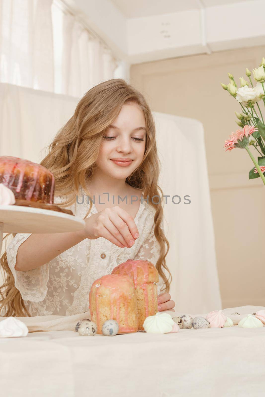 A girl with long hair in a light dress is sitting at the Easter table with cakes, spring flowers and quail eggs. Happy Easter celebration