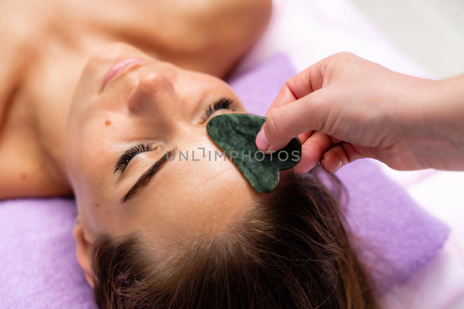 Relaxing massage. European woman getting quartz guache face massage in spa salon, side view.