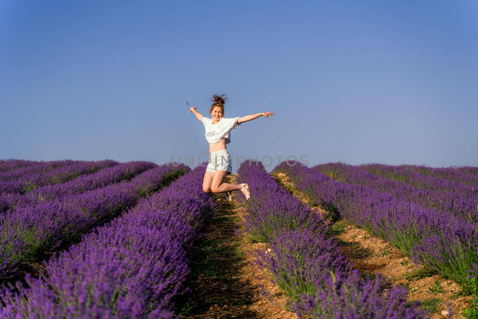 Redhead white woman in white shirt on lavender field smiling, looking at camera and jumping.