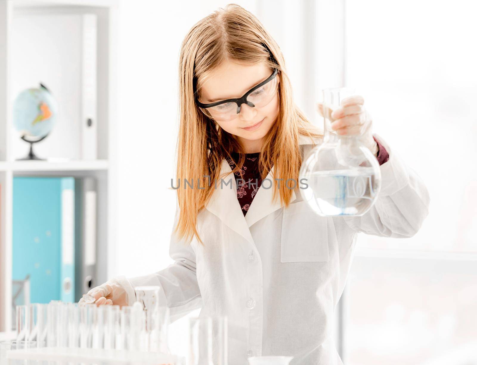 Smart girl doing scientific chemistry experiment wearing protection glasses and holding bottle with substance. Schoolgirl with chemical equipment on school lesson