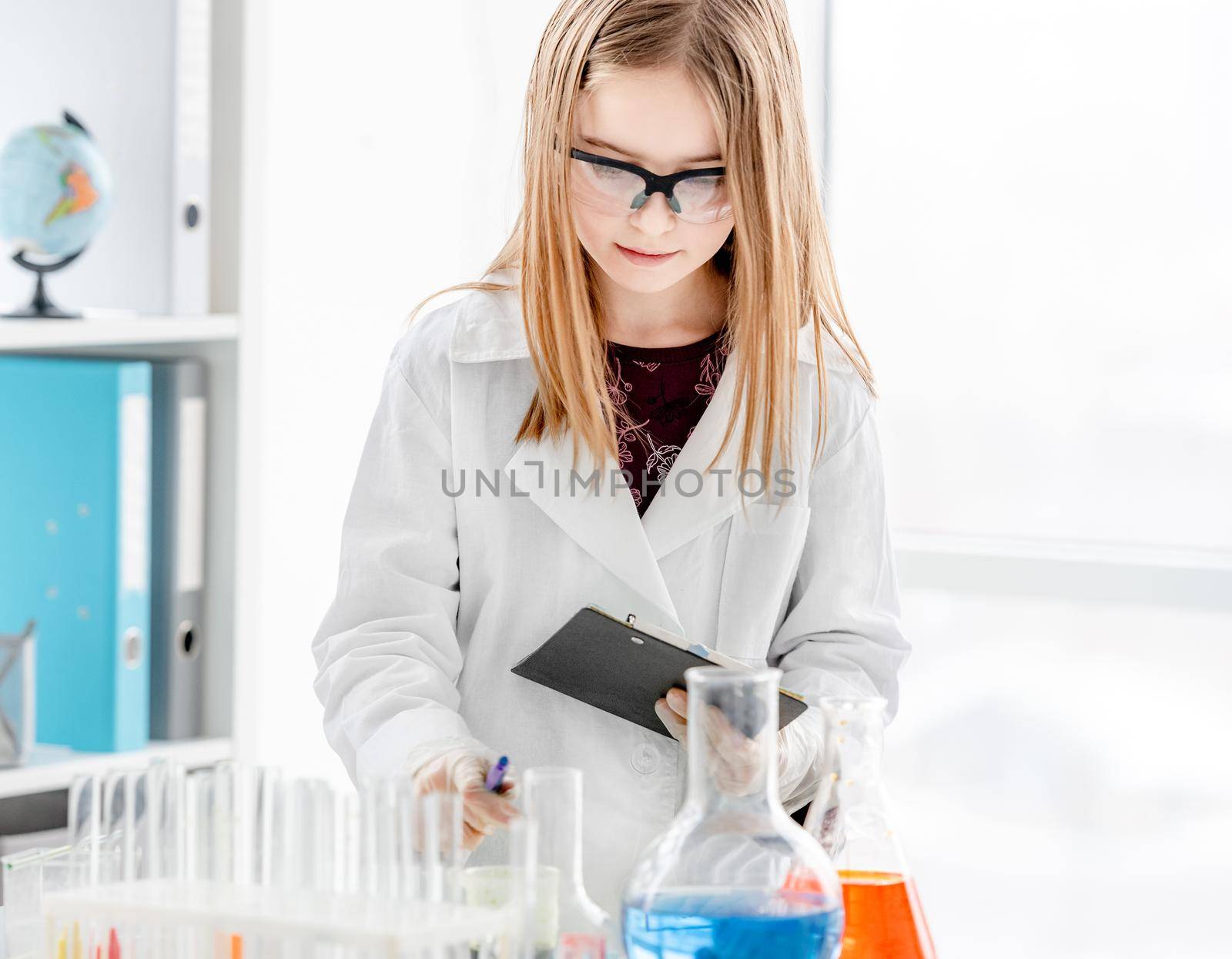 Smart girl during scientific chemistry experiment wearing protection glasses making notes. Schoolgirl with chemical equipment on school lesson