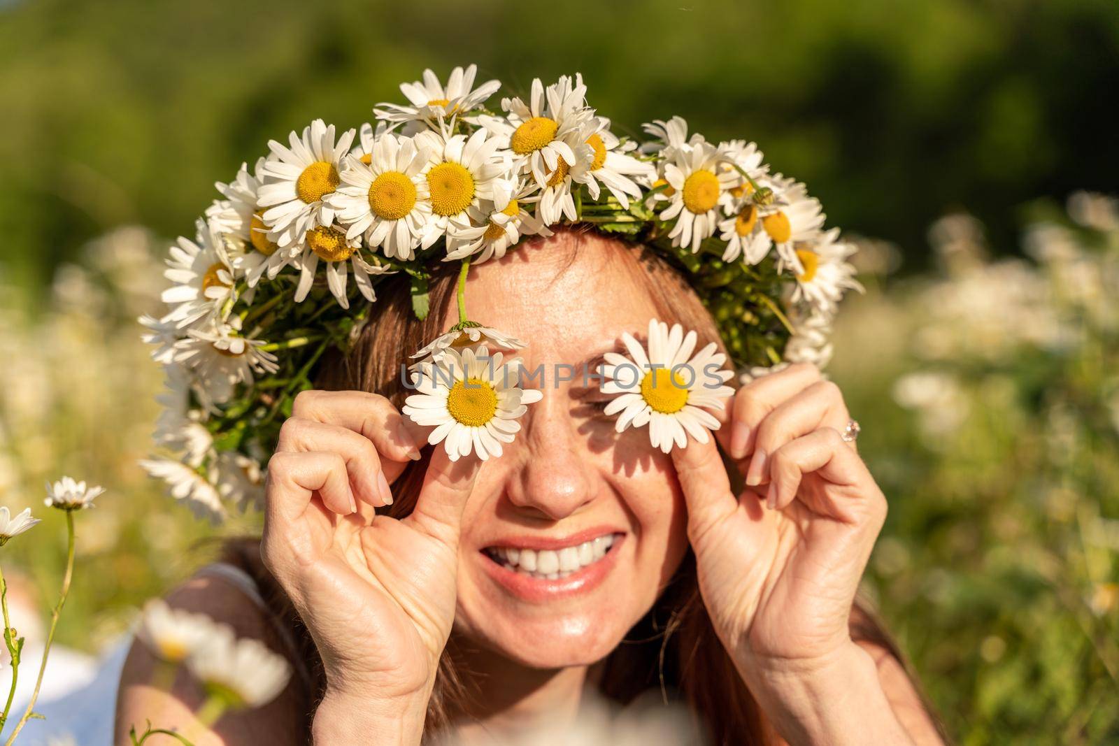 A funny young woman in a wreath of daisies is laughing and holding daisies in front of her eyes. On a large field of daisies against the background of the forest.