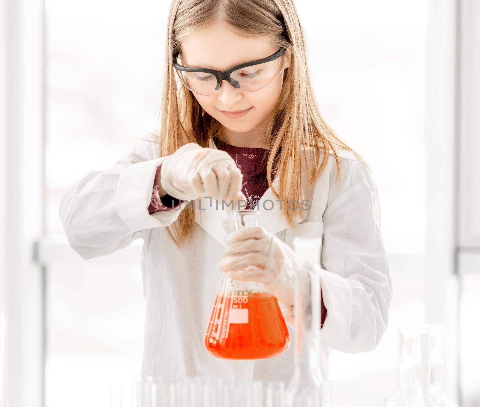 Girl doing scientific chemistry experiment wearing protection glasses and holding tube with orange water. Schoolgirl with equipment and chemical liquids on school lesson