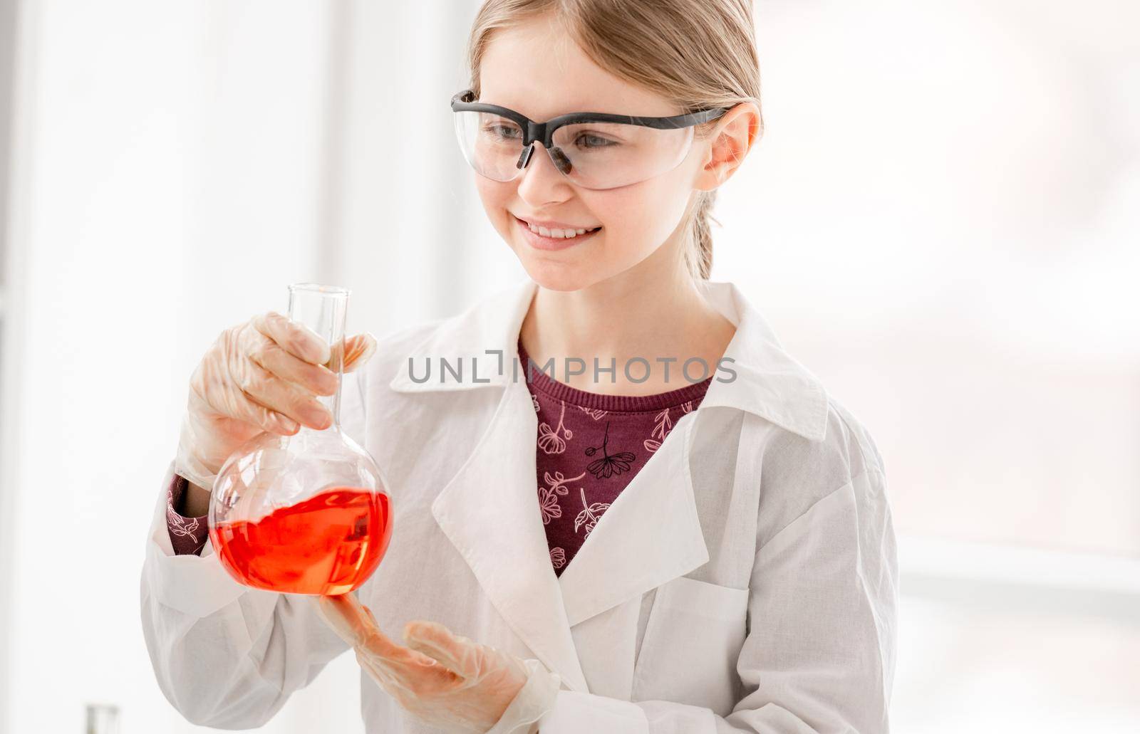 Smart girl during scientific chemistry experiment wearing protection glasses, holding bottle with orange liquid. Schoolgirl with chemical equipment on school lesson portrait