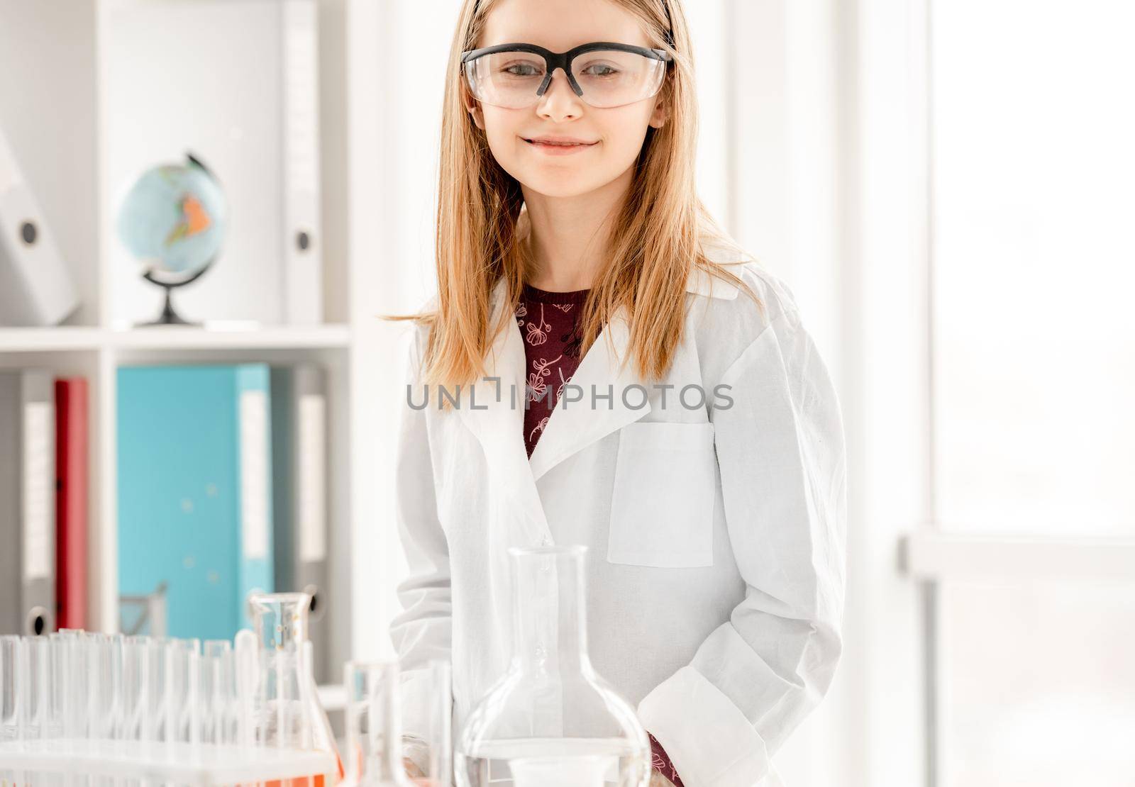 Smart girl during scientific chemistry experiment wearing protection glasses. Schoolgirl with equipment and chemical liquids looking at camera