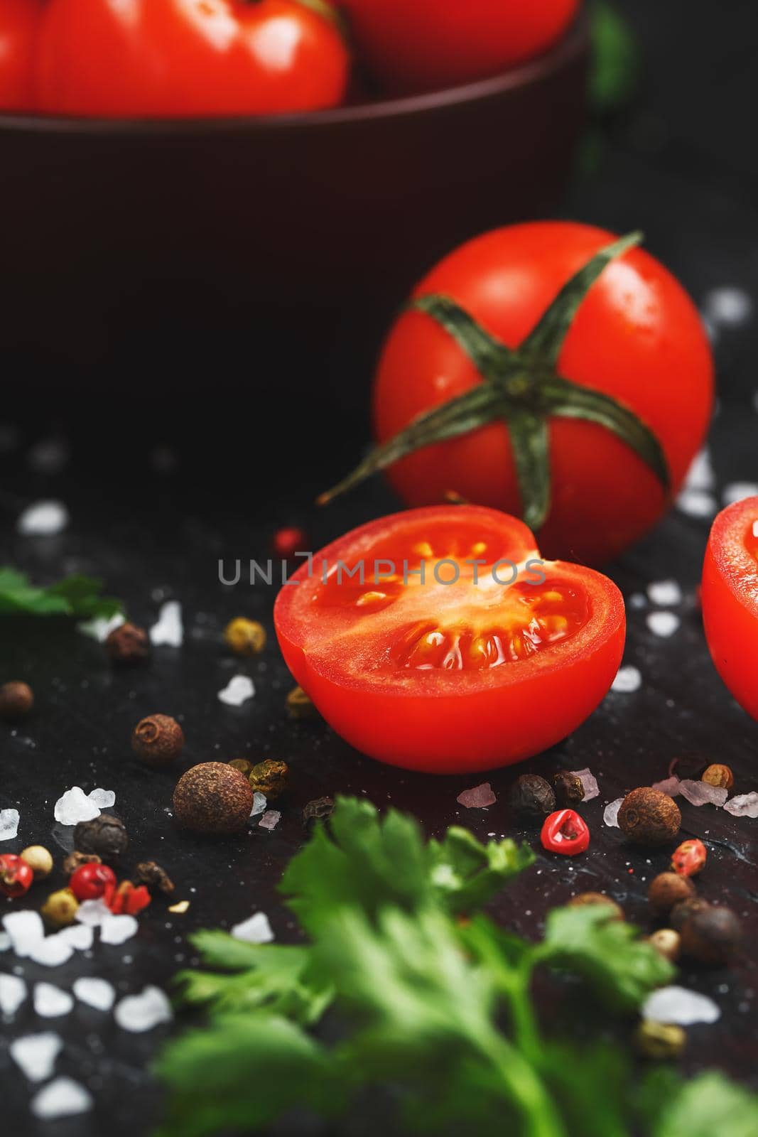 Juicy red cherry tomatoes on a black background with spices, coarse salt and greens. Sliced sweet and ripe tomatoes for salads and as ingredients for cooking. Vertical frame, low contrast