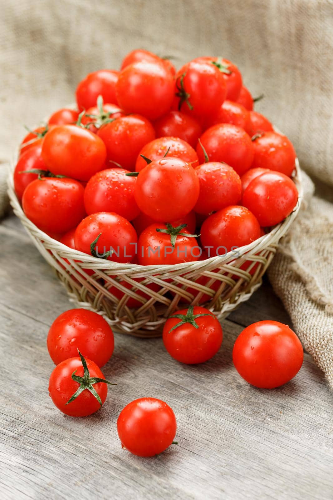 Small red tomatoes in a wicker basket on an old wooden table. Ripe and juicy cherry and burlap cloth, Terevan style country style Vertical frame