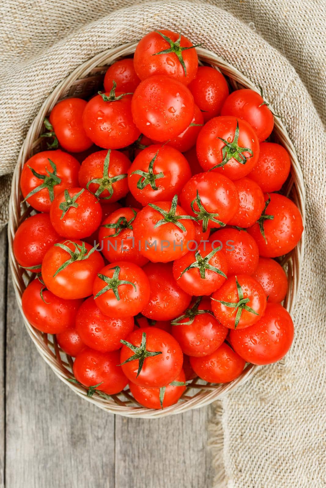 Small red tomatoes in a wicker basket on an old wooden table. Ripe and juicy cherry and burlap cloth, Terevan style country style View from above